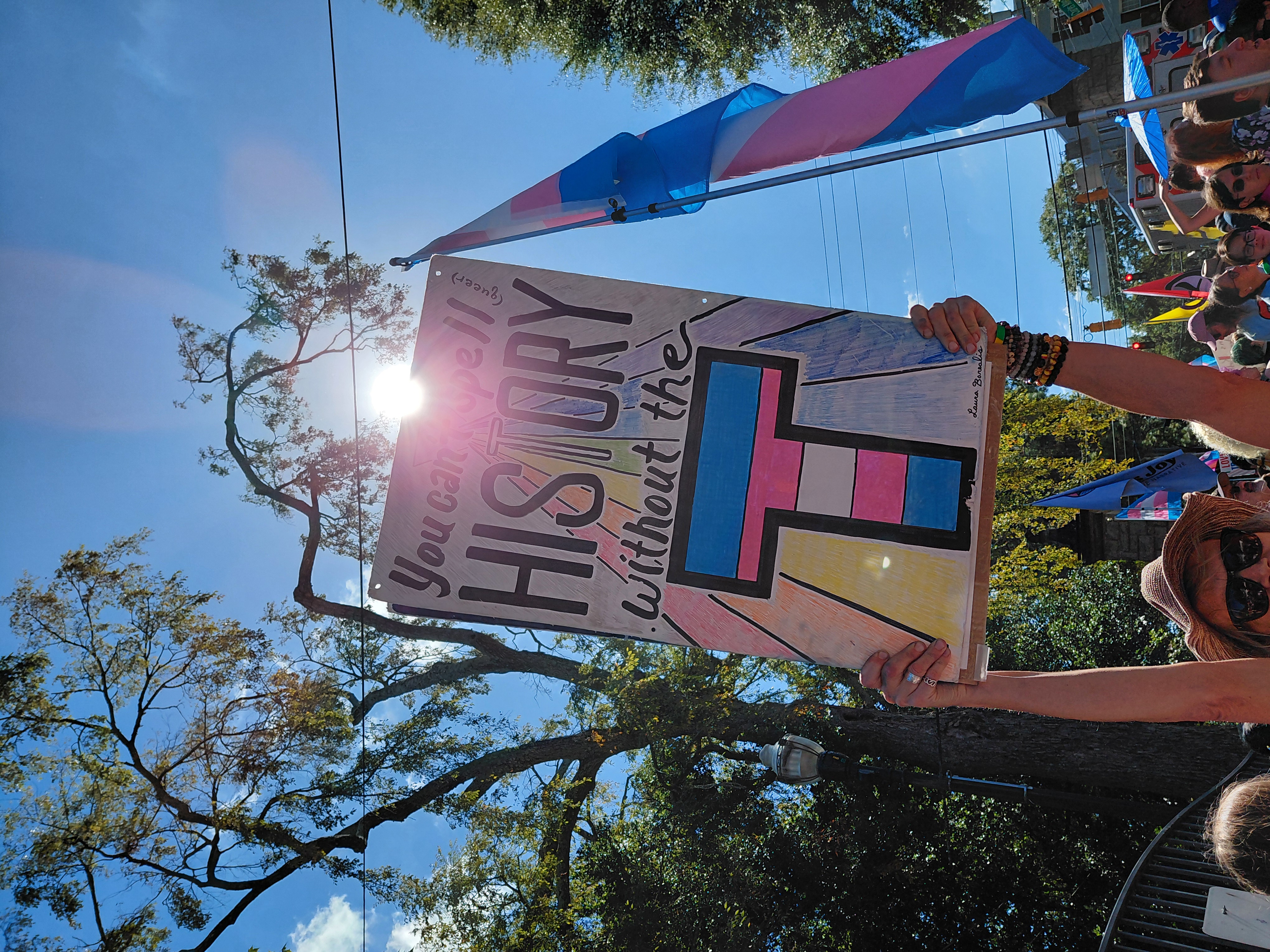 A sign at the Atlanta Pride Trans March reading "You can't spell (queer)
HISTORY without the T" with the "T" in the colors of the trans flag.
The sign is illuminated from behind by the Sun.